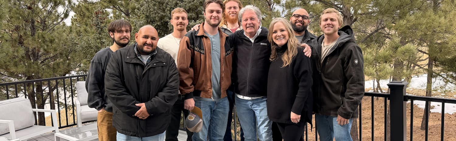 A group of nine people, dressed in casual jackets and standing closely together, smile for a photo outdoors on a deck with trees and a hint of snow in the background.