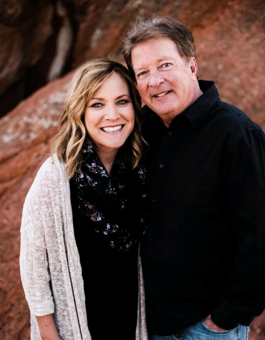 A smiling woman and man stand close together outdoors in front of large reddish rocks. The woman has blonde hair and wears a light sweater and dark scarf, while the man wears a black shirt and jeans.