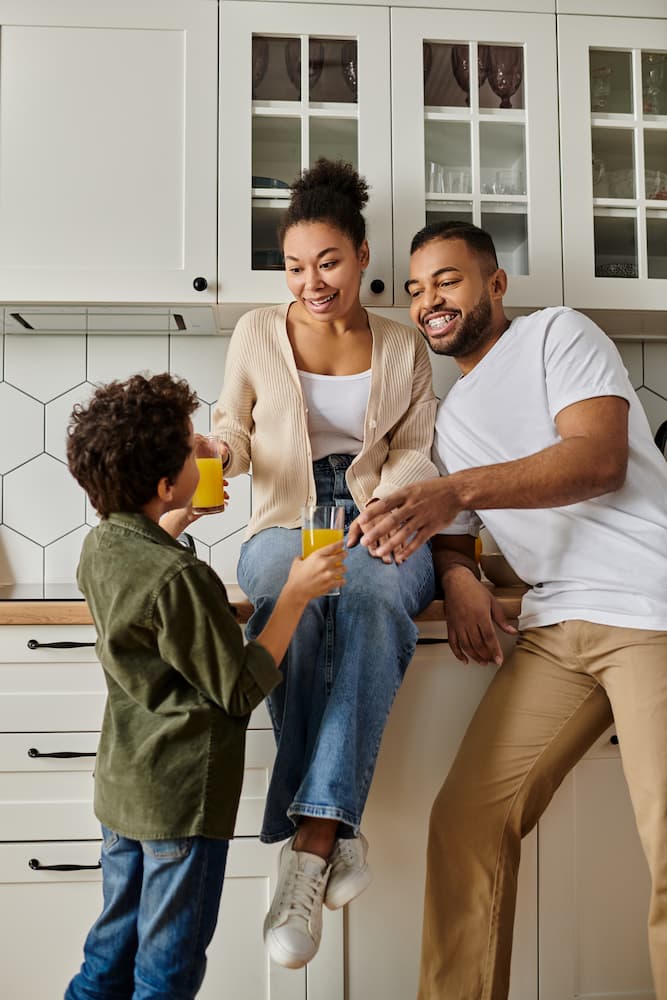 A smiling family of three enjoys glasses of orange juice together in a bright kitchen, with the parents sitting on the counter and their child standing, all raising their glasses in a cheerful toast.