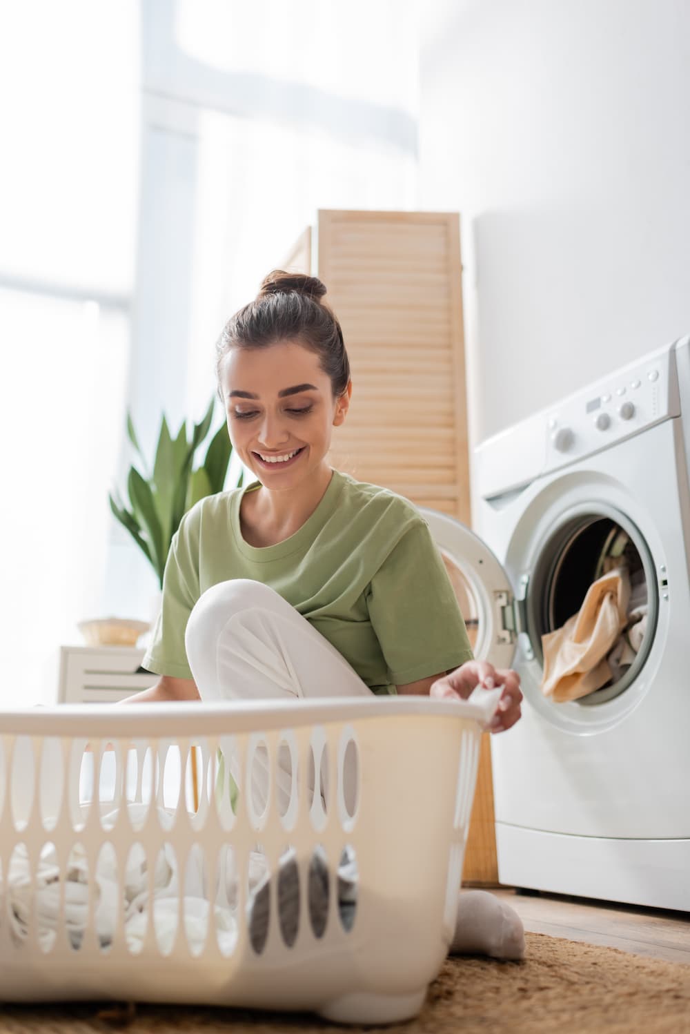 A smiling woman sits on the floor in front of a washing machine, sorting laundry into a white basket. The room is bright with natural light and a green plant is visible in the background.