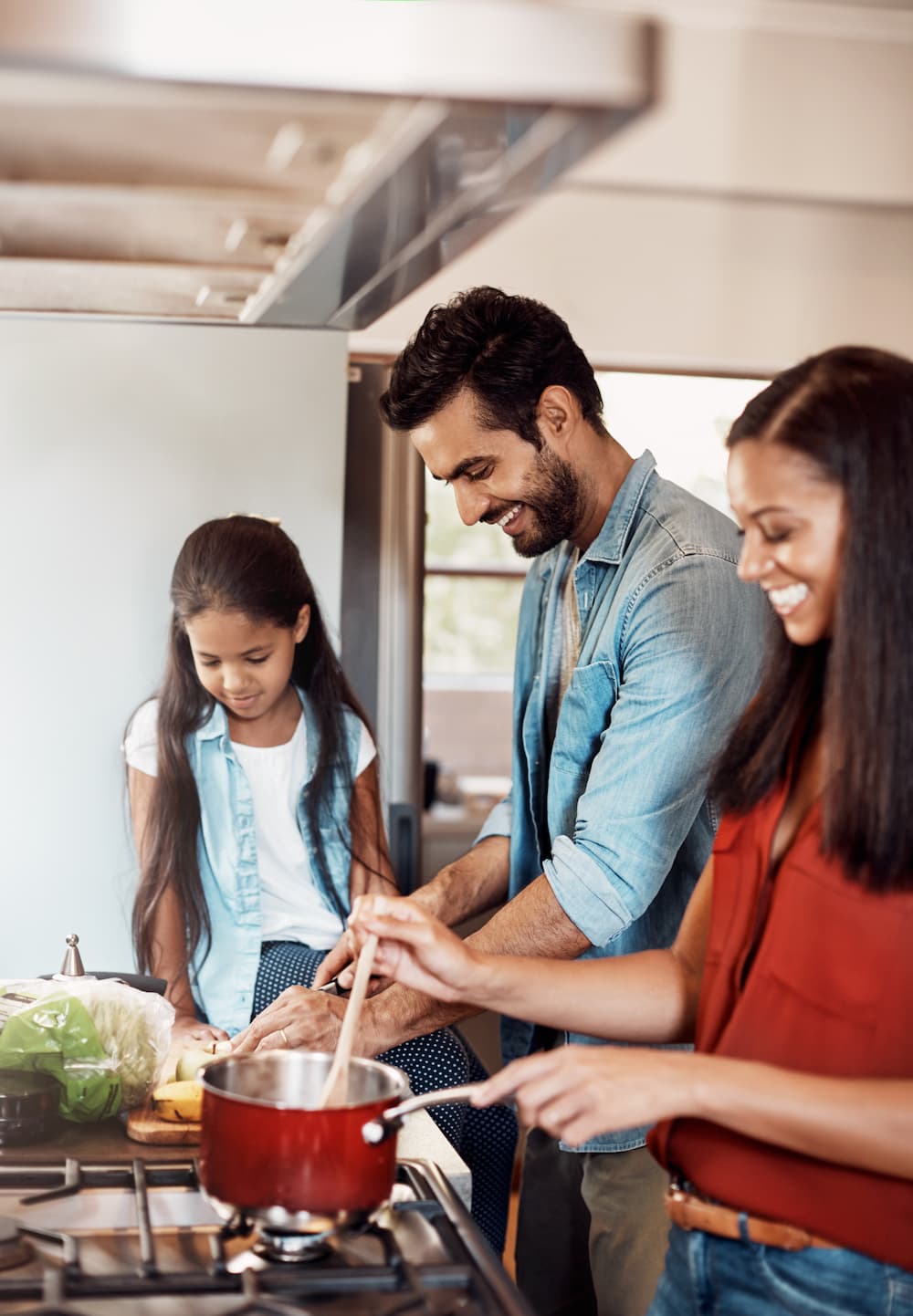 A smiling family stands in a kitchen cooking together. An adult stirs a pot on the stove while another adult and a young girl assist, surrounded by fresh vegetables and ingredients on the counter.