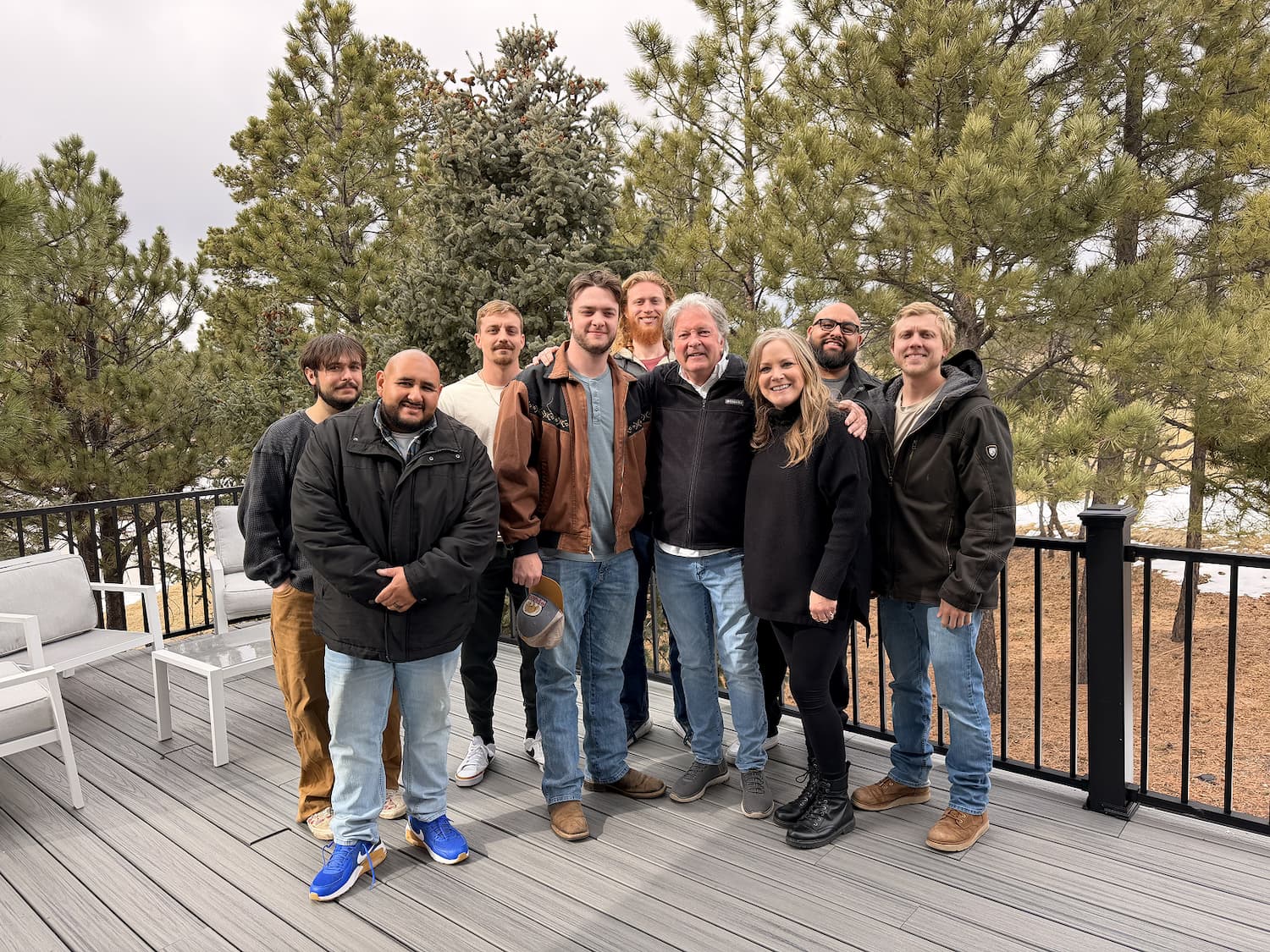A group of nine adults, dressed in casual winter clothing, stand close together and smile on a deck with pine trees and a cloudy sky in the background. Two white chairs are on the left.